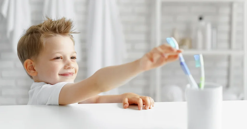 Smilende ung dreng rækker ud efter en tandbørste fra en kop. Mundhygiejne børn